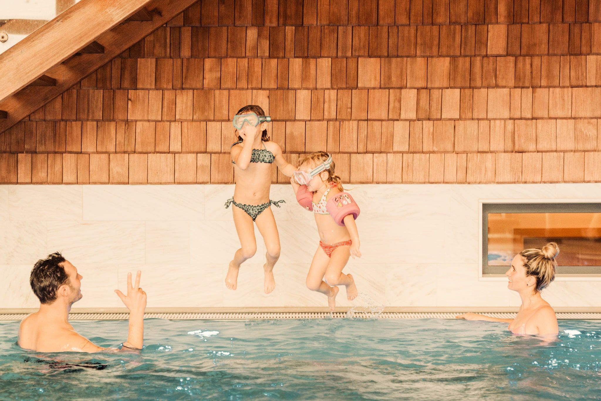 Two girls jump into the water while adults relax in the pool.