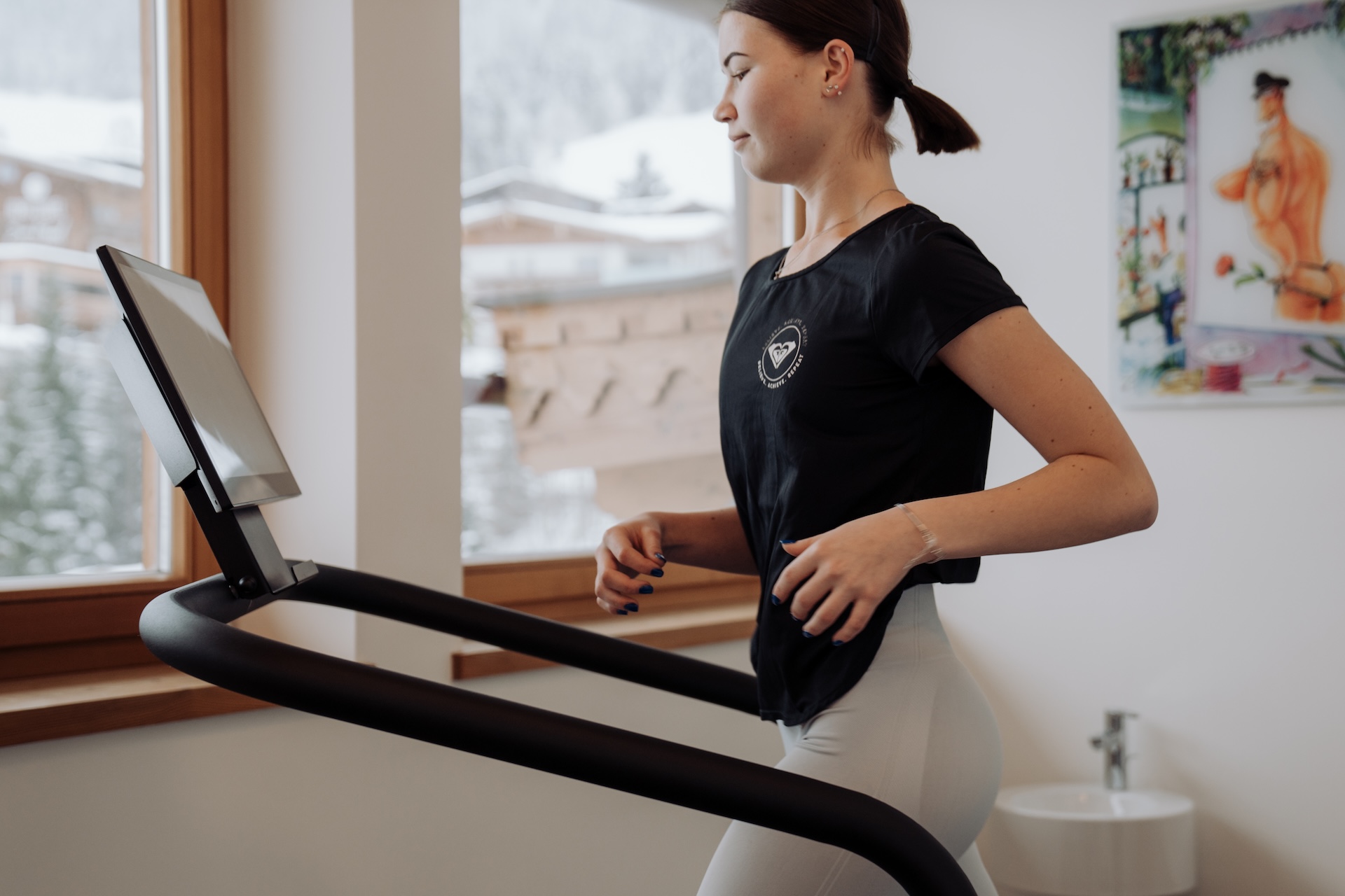 A woman is running on a treadmill with a view of winter scenery outside, possibly at the Glemmtalerhof in Hinterglemm.