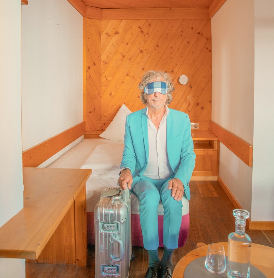Ein Mann in aquafarbenem Anzug sitzt im Glemmtalerhof mit Koffer und blickt nachdenklich ins Leere.