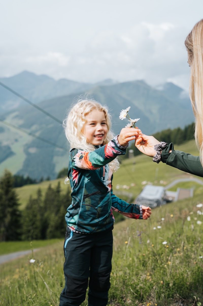 Ein Kind mit lockigem Haar reicht einer Person eine Blume, umgeben von den schönen Bergen des Glemmtals.