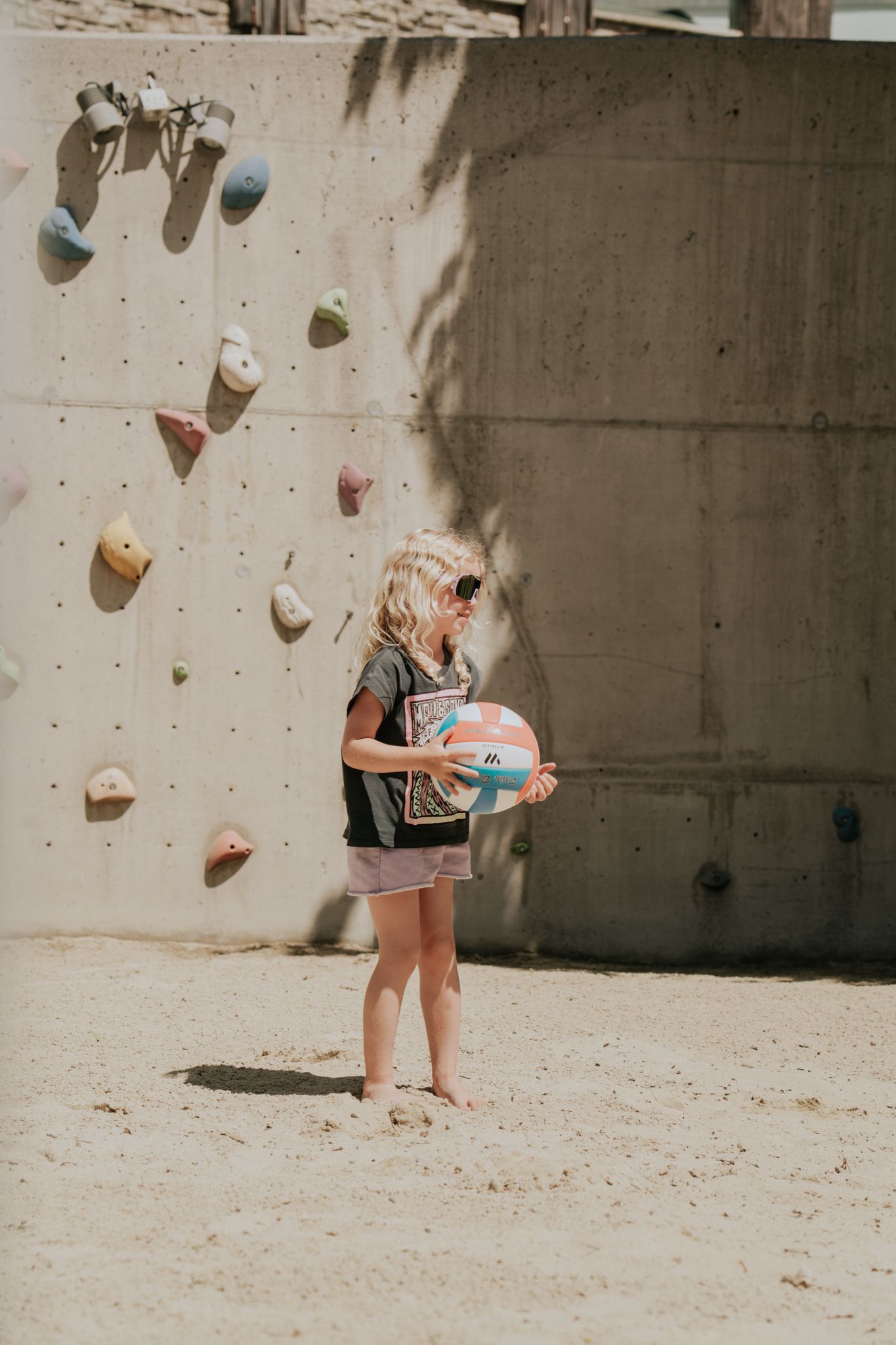 A girl in the sand with a ball, climbing wall behind her. Glemmtal adventure.