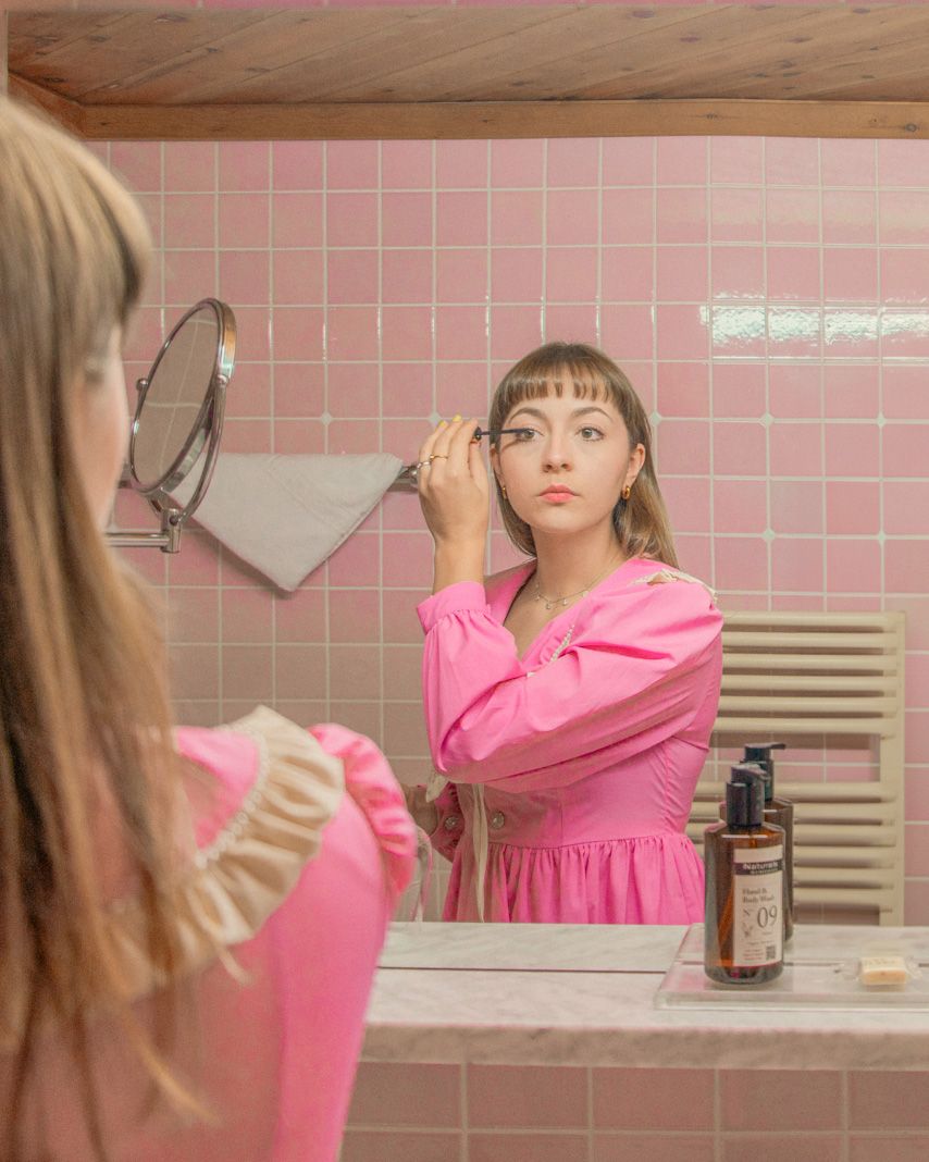 A woman is applying makeup in a pastel-colored bathroom. The Glemmtalerhof might be nearby.
