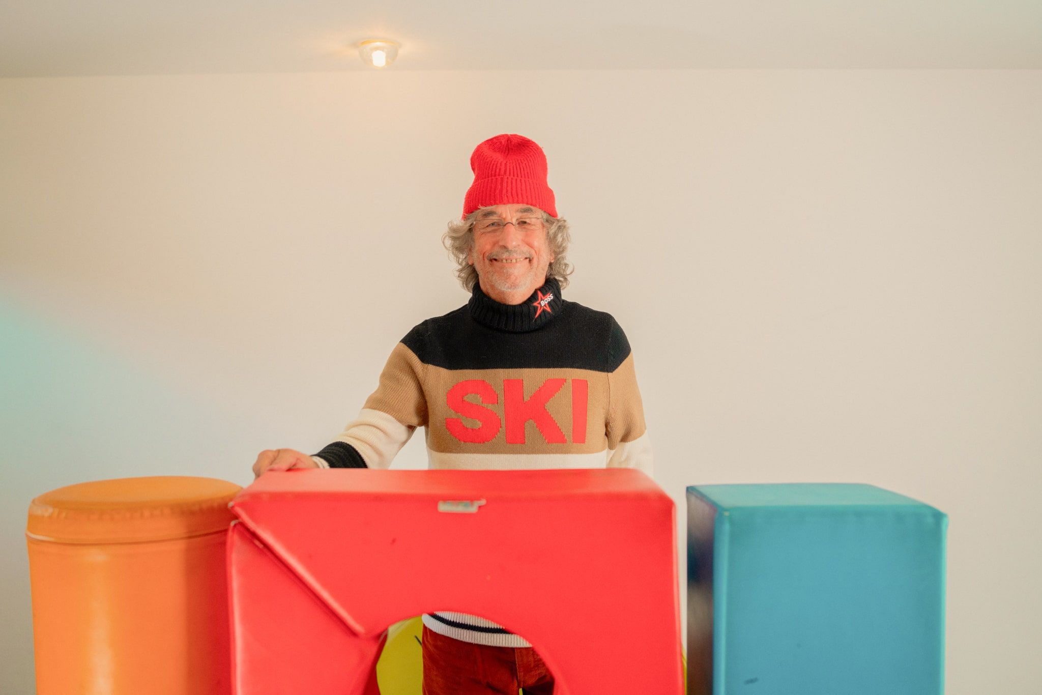 Ein lächelnder Mann mit einem SKI-Pullover steht vor bunten Objekten im Glemmtal.