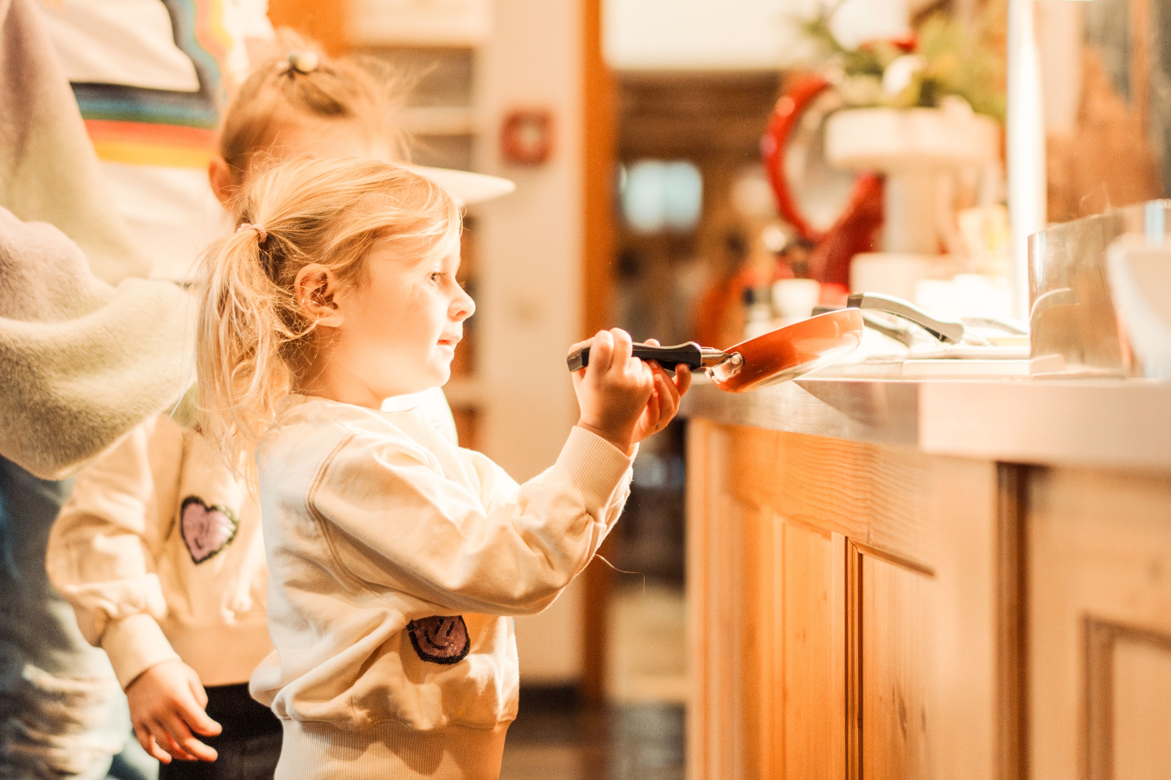 A little girl in the kitchen with a pan, cheerful and curious.