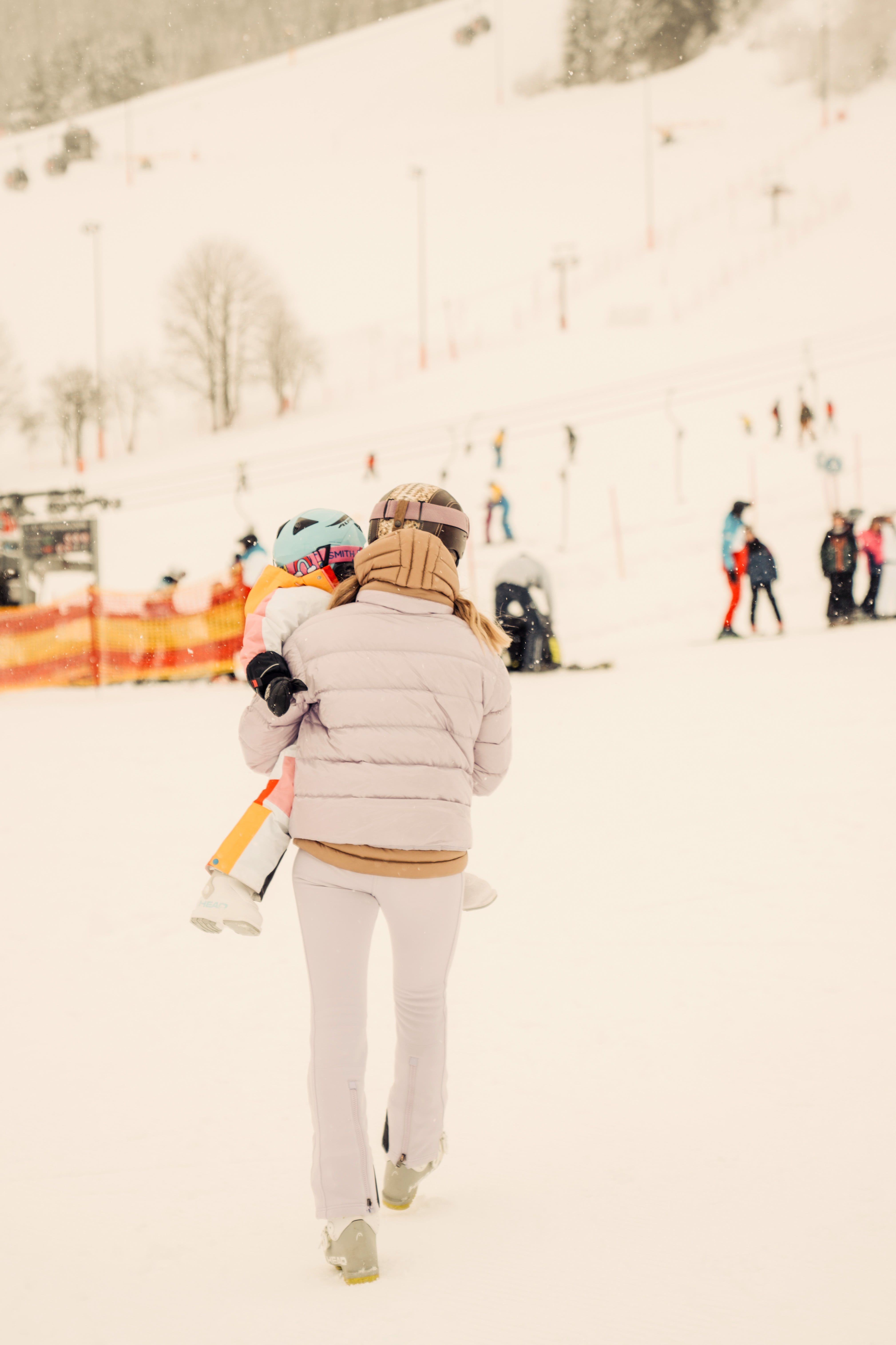 Eine person trägt ein kind im Glemmtal beim Skifahren. Viel Schnee und fröhliche Menschen sind sichtbar.